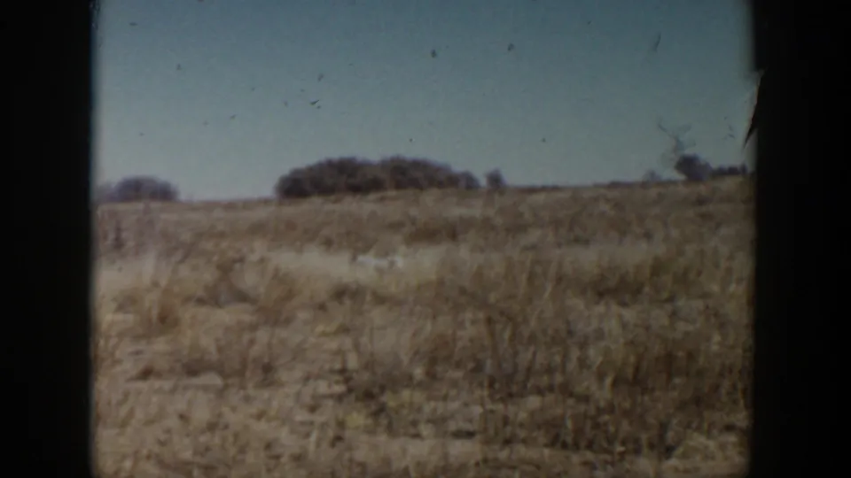 1957 Lubbock, Texas 8mm view of dry prairie terrain, distant shrub clusters, and clear sky from a moving vehicle.