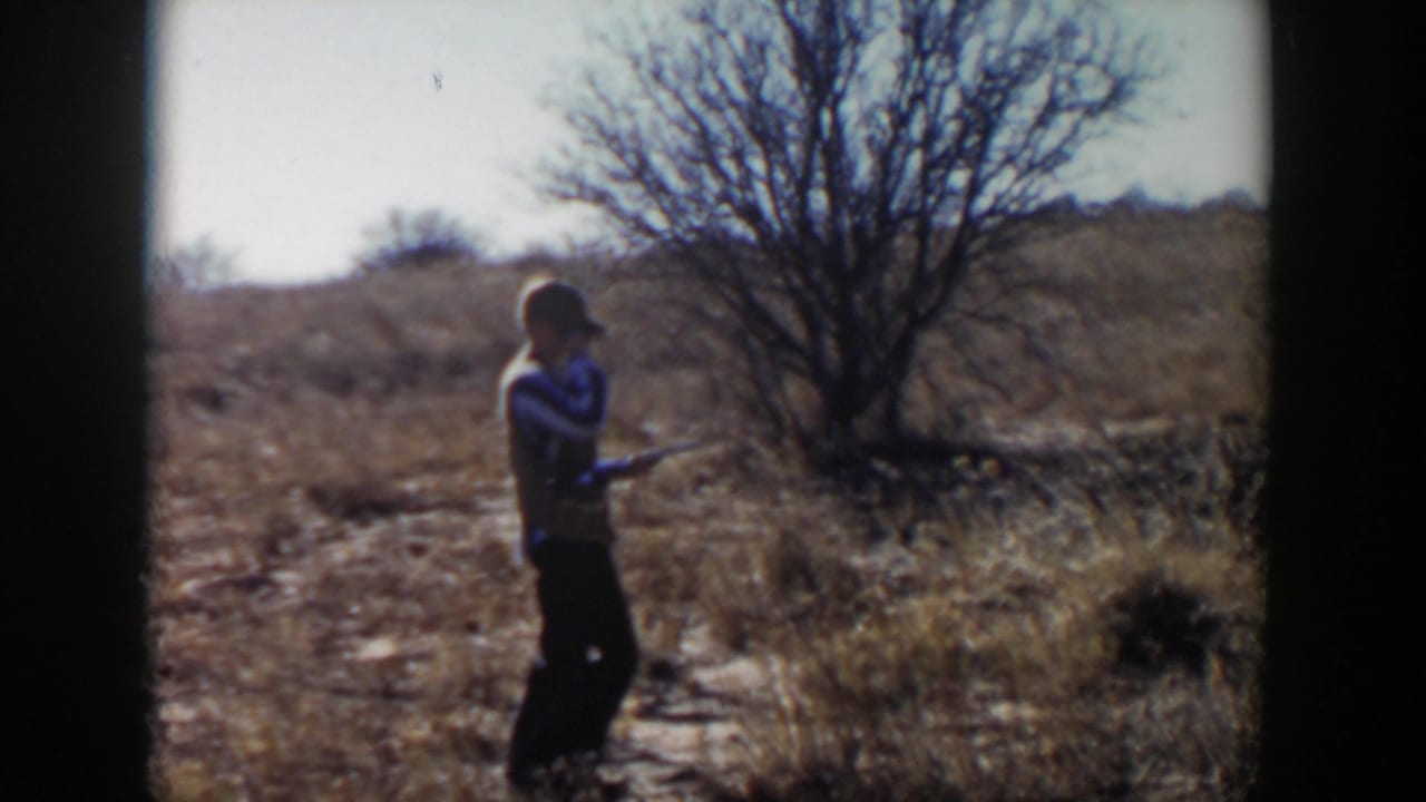Man in a cap holding a rifle in a dry field near a leafless tree.