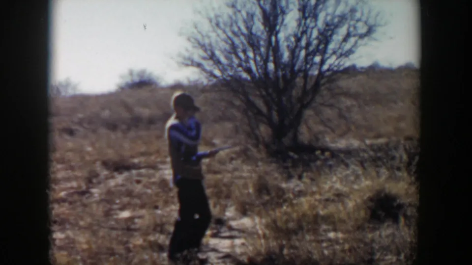 1957 Lubbock-area home-movie frame of a man with a rifle standing in dry grass near scrub and a bare tree.