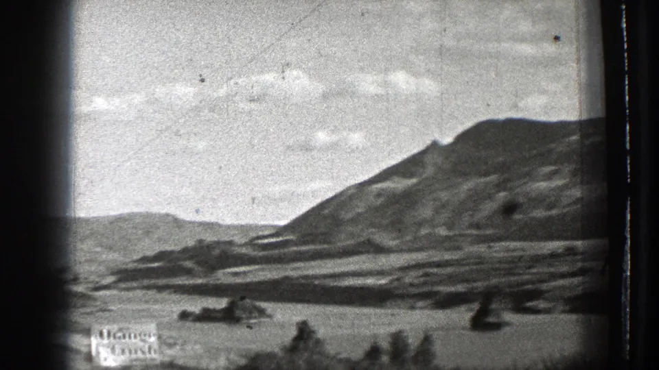 1939 8mm home-movie frame showing rugged hills, pale sky, and a rocky water or dry basin area seen through dark film-gate edges.