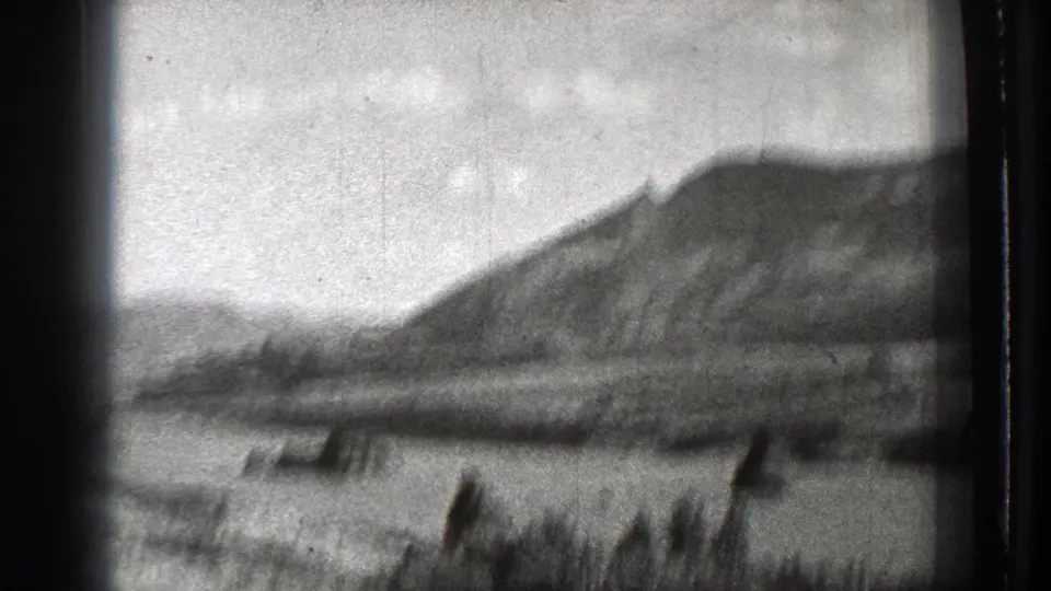 Soft 1939 8mm frame of a mountainous horizon, broad basin or water area, and blurred foreground vegetation from a moving viewpoint.