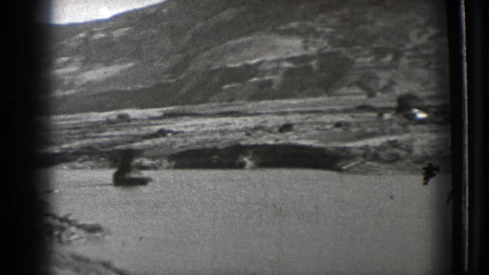 1939 8mm frame showing a wide mountain slope, open basin or water edge, rocky foreground, and visible film scratches.