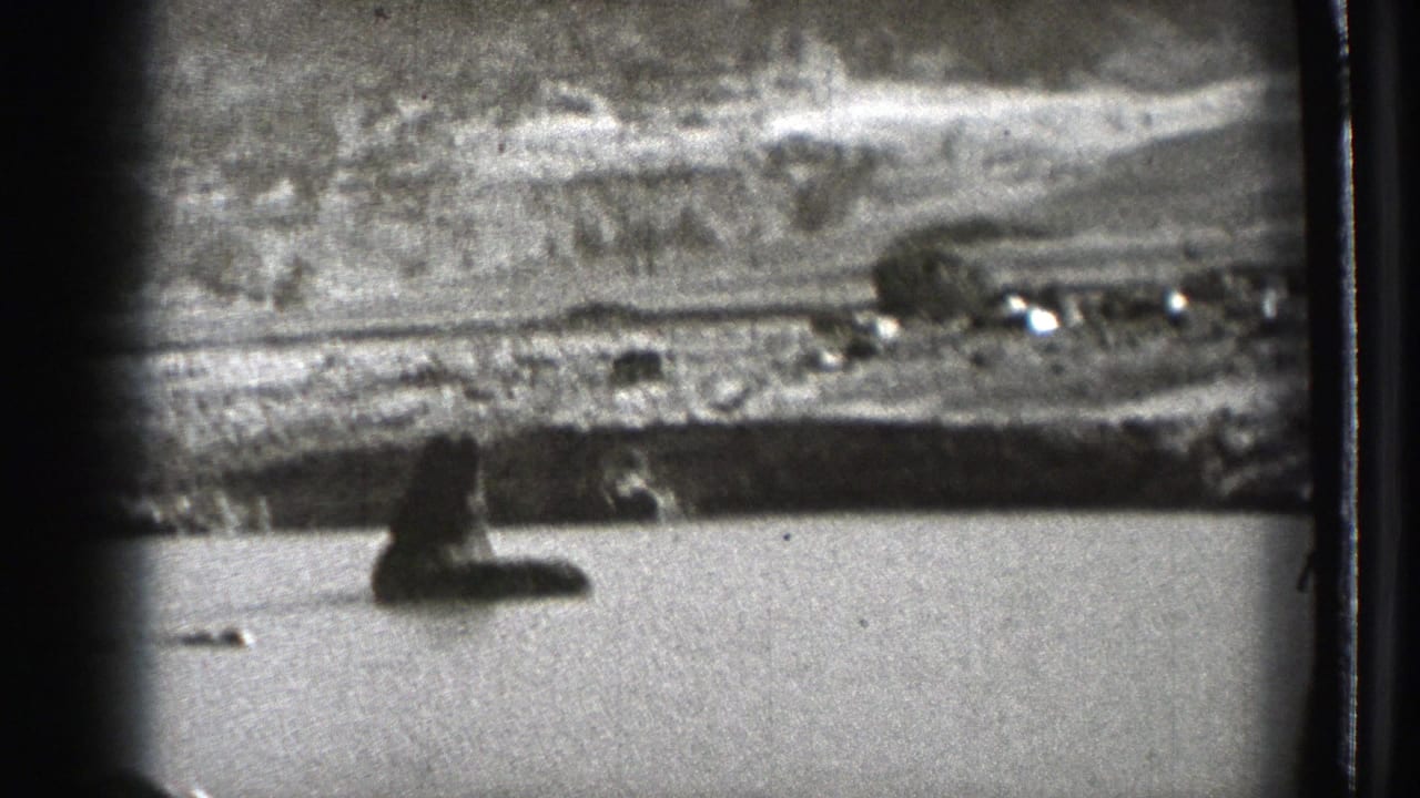 1939 Washington 8mm frame showing a lake, distant hills, shoreline structures, and dark foreground shapes from a moving viewpoint.
