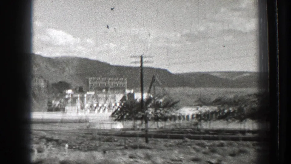 1939 Washington home-movie frame passing a dam or industrial site, with a roadside sign, utility pole, hillside, and brush visible.
