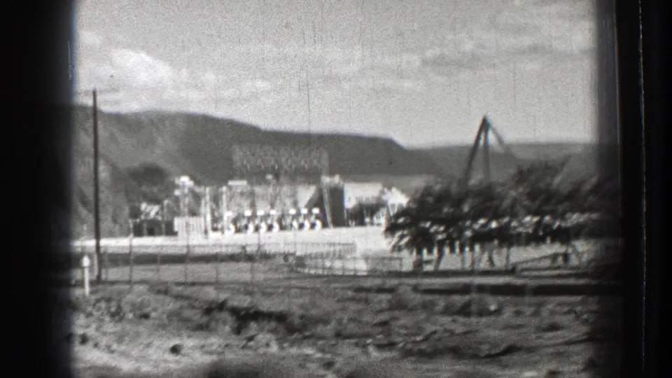 Late-1930s Washington road-trip view of a distant dam or utility complex, rough foreground, utility poles, hills, and an angled structure.