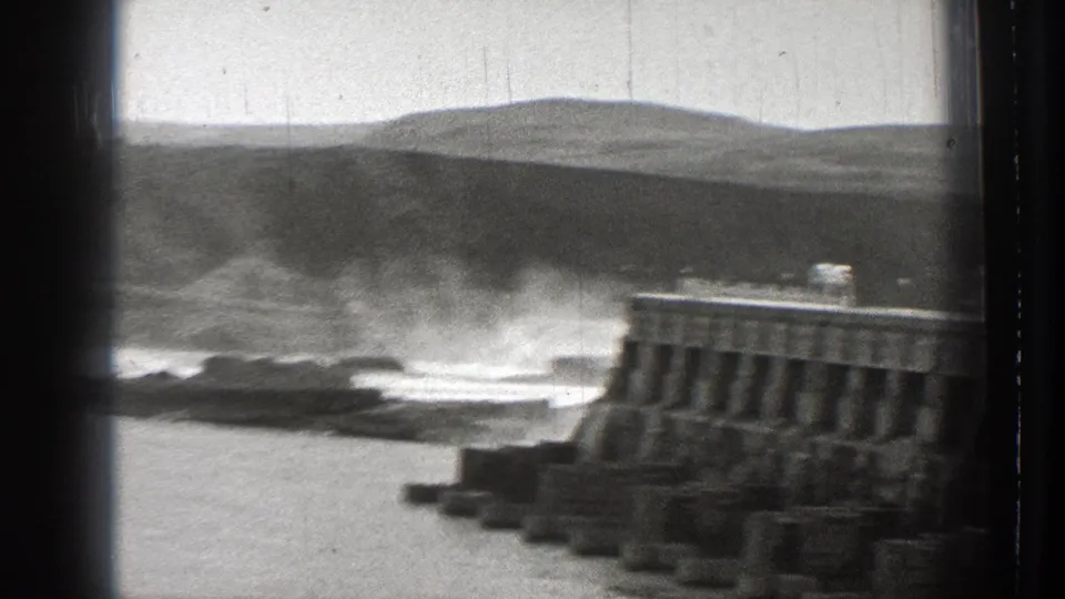 Home-movie frame of the Columbia River dam with churning water, concrete supports, and a low hilly background.