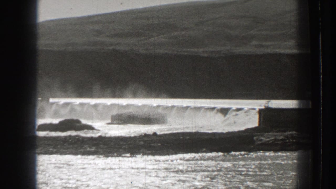 1939 Washington 8mm frame showing a low dam releasing water, with mist, rocks, reflective water, and a dark hillside in the background.