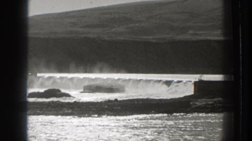 1939 Washington 8mm frame showing a low dam releasing water, with mist, rocks, reflective water, and a dark hillside in the background.