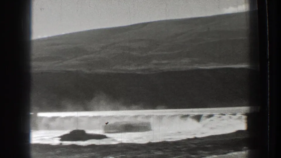 1939 Washington 8mm frame of water cascading over a dam, seen across a rocky shoreline with a dark hillside behind the spillway.