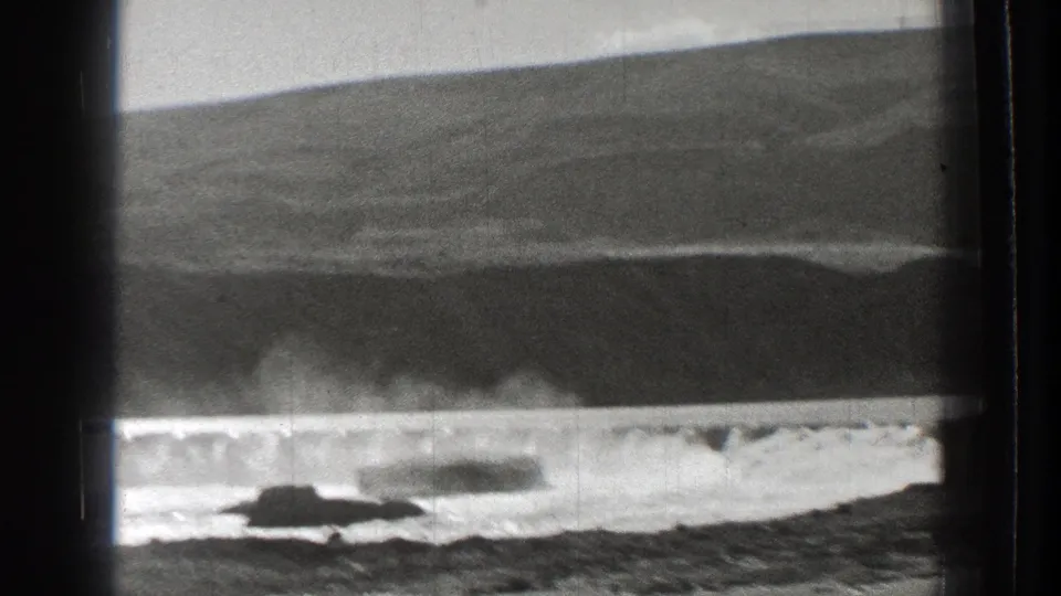 1939 Washington 8mm frame showing foaming water beneath a low dam, with a dark sloped hillside filling much of the background.
