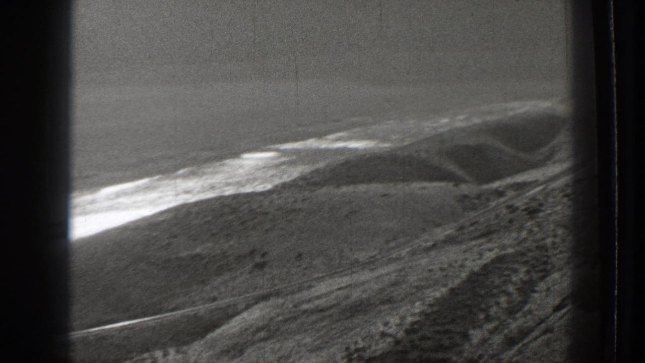 Aerial view across sloping hills, pale shoreline, water, and distant ridges.