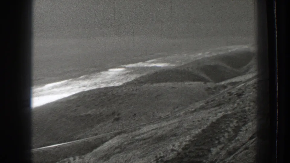 1939 Washington aerial frame with sloped terrain, a bright shoreline band, water beyond, and hazy ridges under a pale sky.