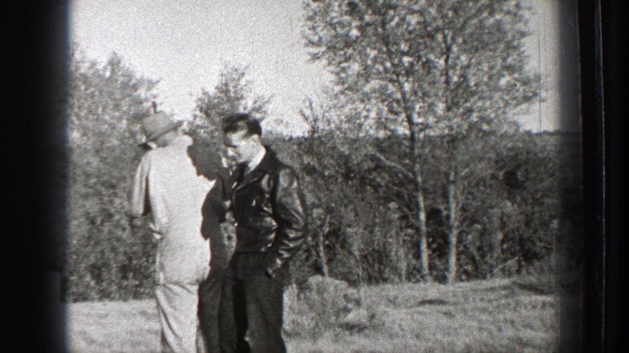 Two adult men stand in a grassy field near trees, one in a hat and one in a dark jacket.