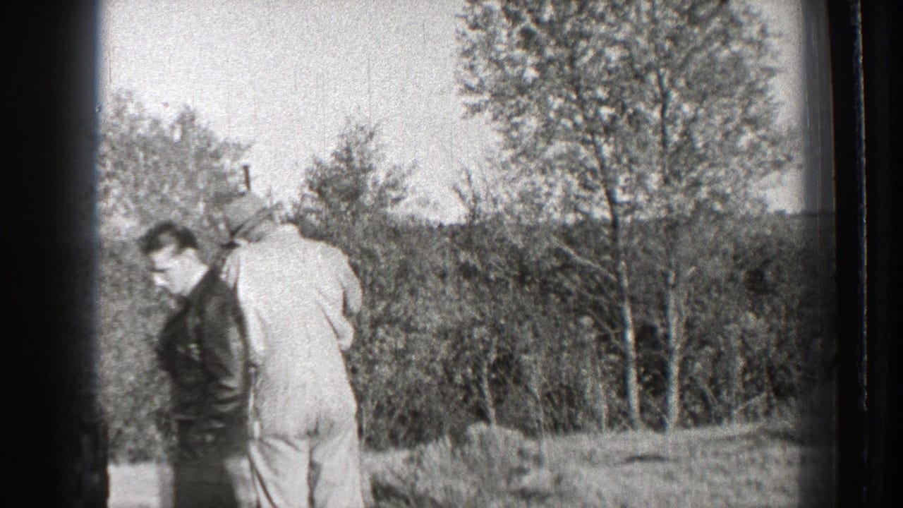 Two men stand close together outdoors, partly blurred, with trees behind them.