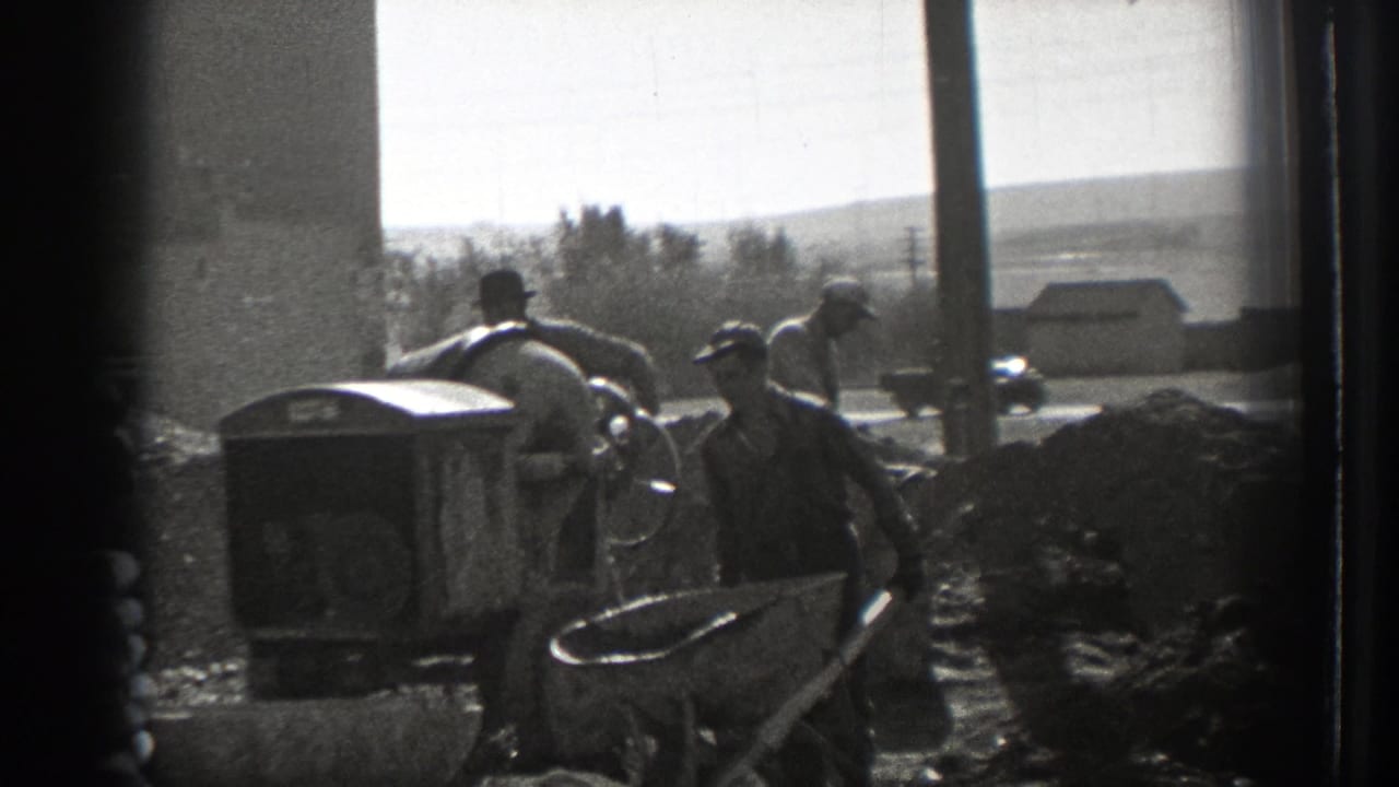 Workers stand near a concrete mixer and wheelbarrows at an outdoor construction site.