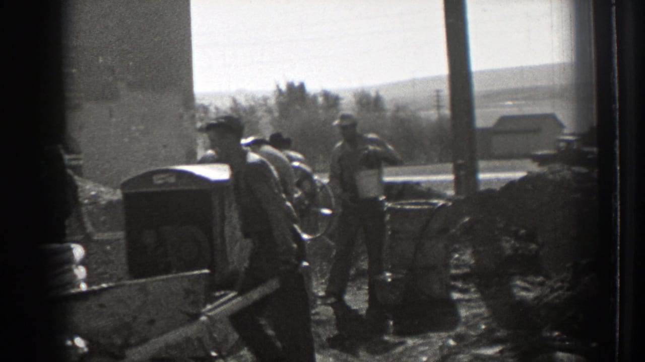 Men work around a concrete mixer with wheelbarrows and material piles nearby.