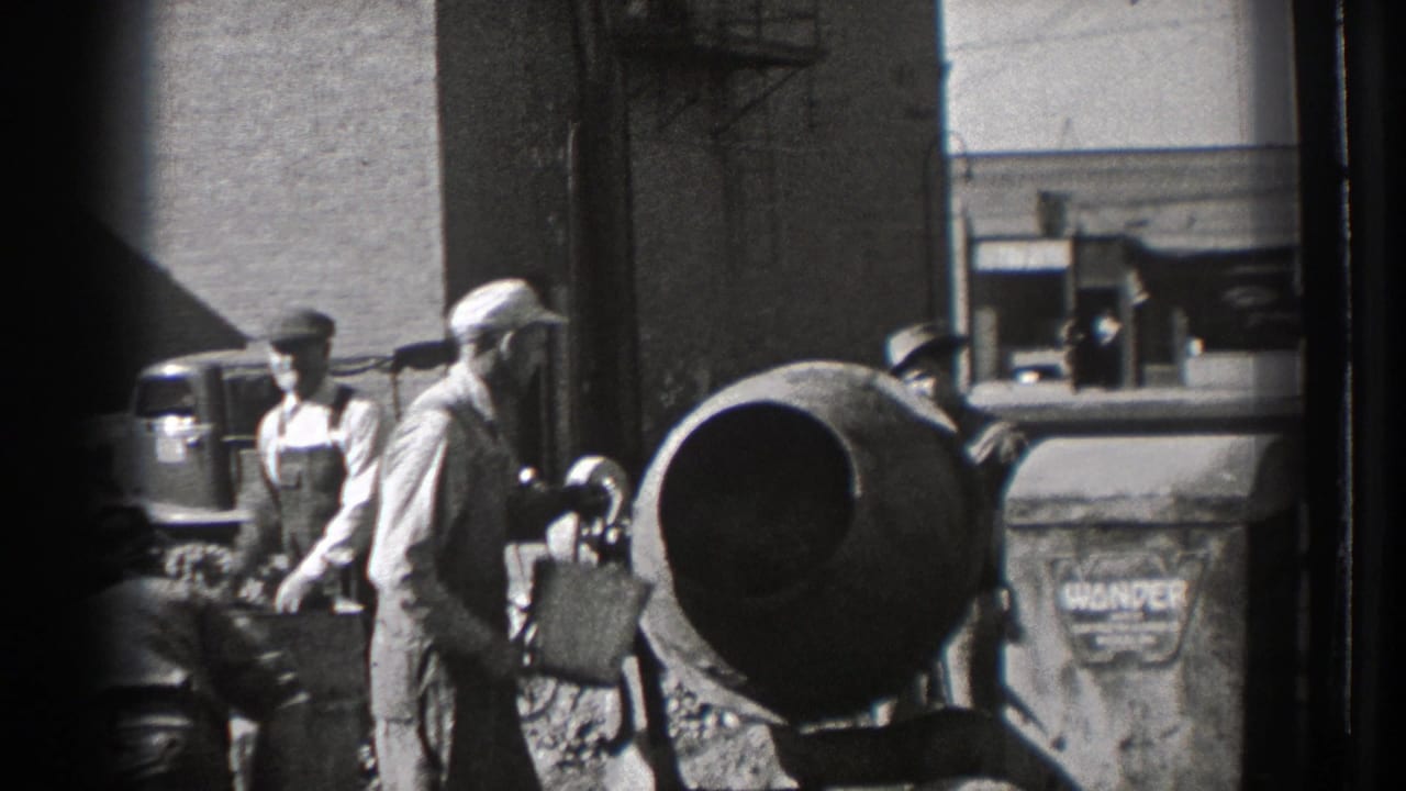 Workers stand near a cement mixer beside a brick industrial building.