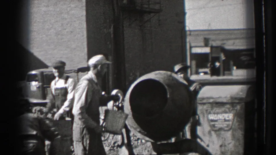 Washington 1939 worksite view with men in caps and overalls near a cement mixer, aggregate piles, a brick wall, and a vintage automobile.