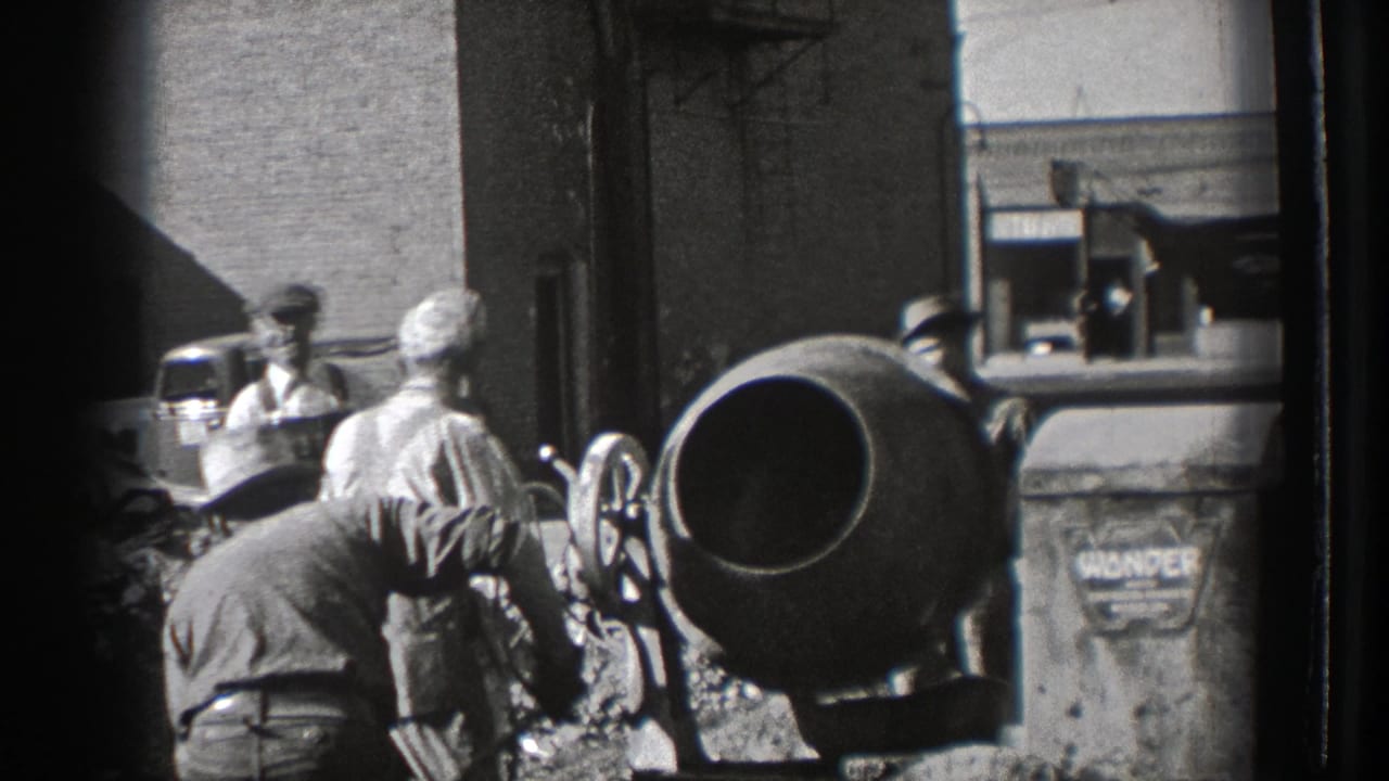 Men work around a cement mixer and wheel-handled equipment near a brick wall.