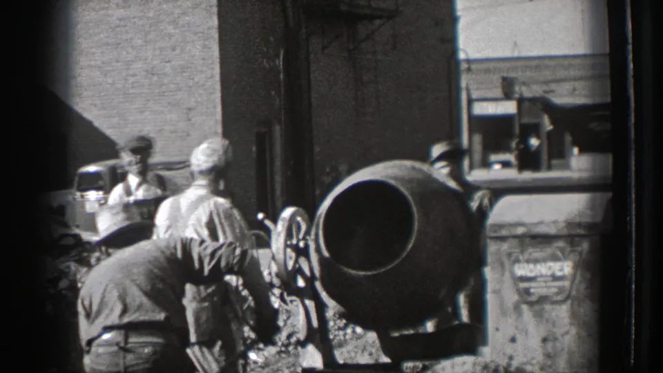 Workers at the 1939 Washington site bend and stand around a cement mixer, with a brick building, window, automobile, and sign-like marking in view.
