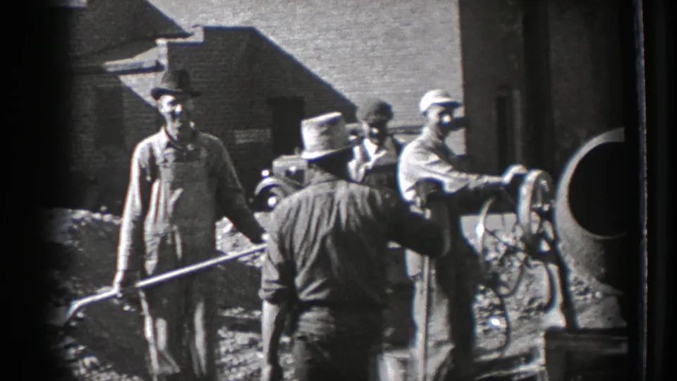 Washington 1939 construction workers in caps and overalls use shovels near a cement mixer and material piles beside an industrial brick structure.