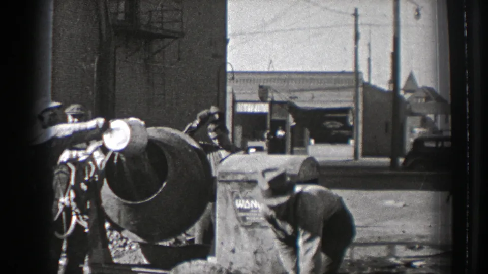 Washington 1939 worksite frame showing a man bending near a barrel, another by the cement mixer, and street buildings beyond.