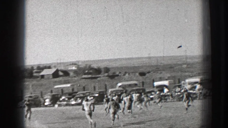 Washington 1939 home-movie frame showing people running across a field near parked cars, houses, and low hills.