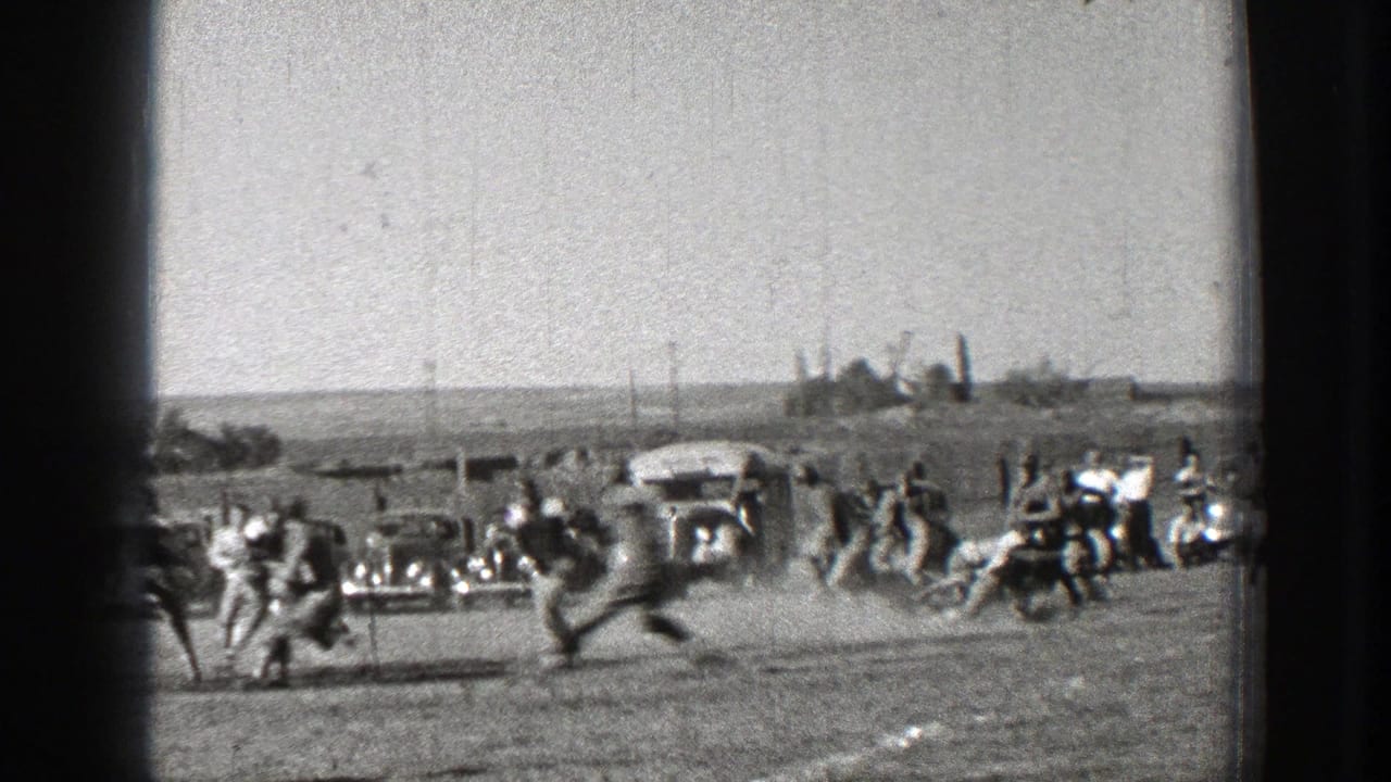 Football players run across a dusty field with spectators and parked cars behind them.