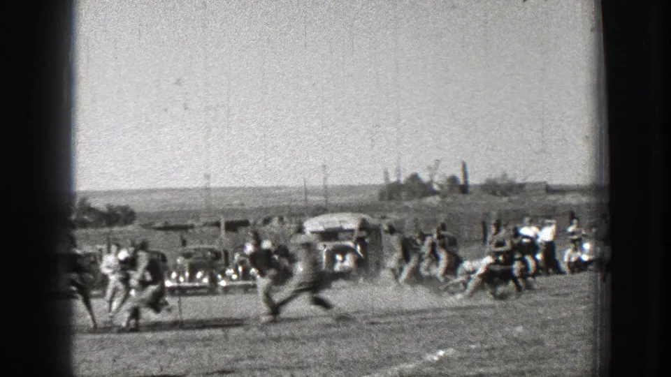 Washington 1939 home-movie frame showing football players in motion on a dusty field, with spectators and period cars along the sideline.