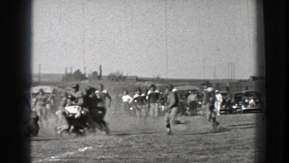 A 1939 Washington football game frame showing players spread across the field, dust in the air, and spectators near parked cars.