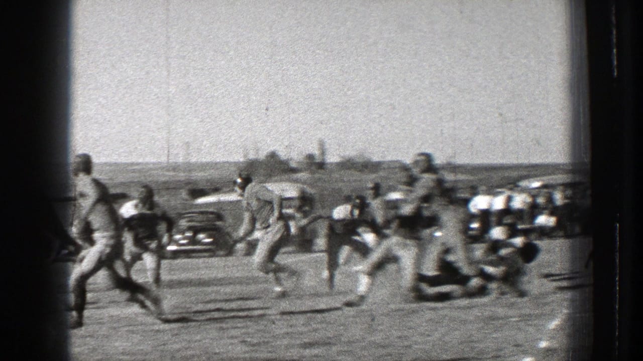 Football players run during a play with cars and spectators in the background.