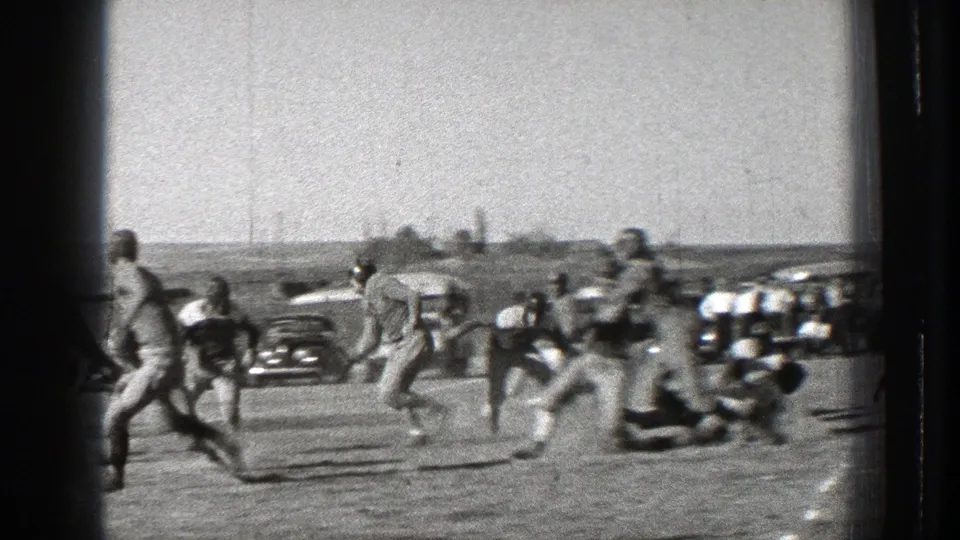 Players in football uniforms move across the field during a play, with vintage automobiles visible behind them.