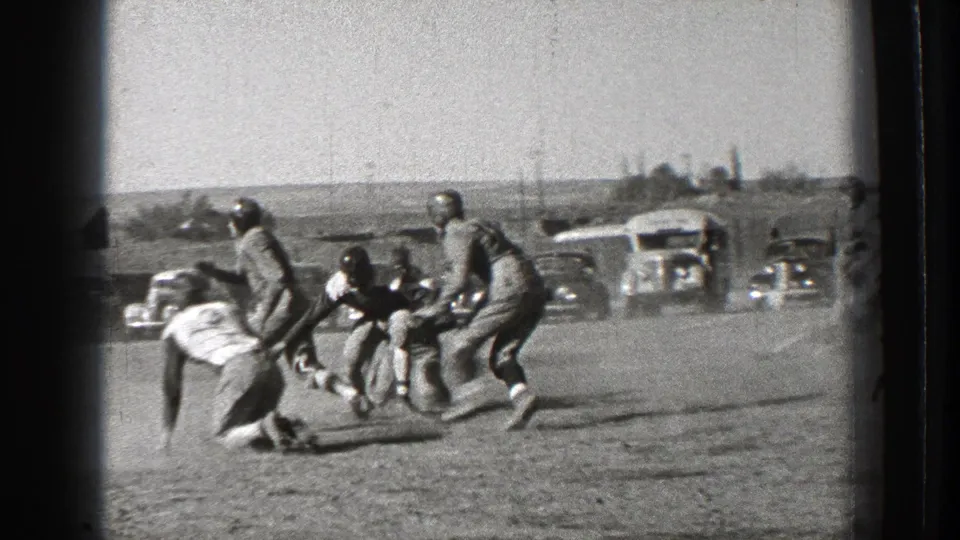 A football tackle unfolds on the field, with players clustered together and vintage cars beyond the action.