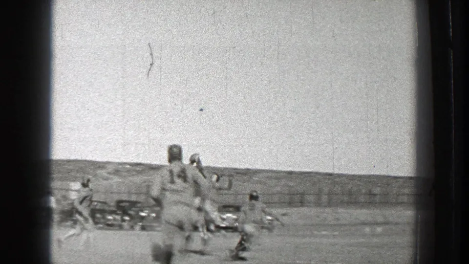 A late-game view shows football players moving downfield across an open Washington field, with cars and fencing beyond.