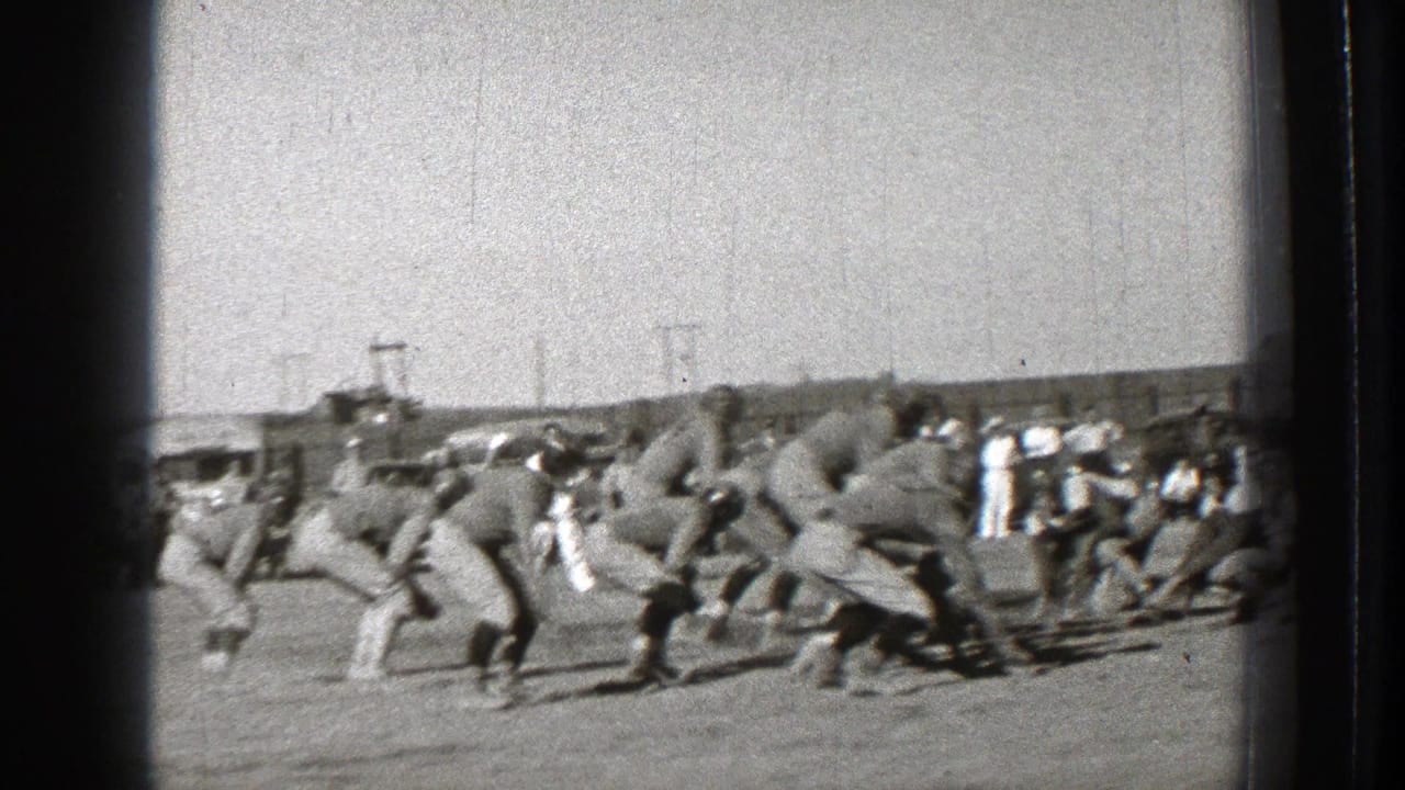 In 1939 Washington footage, helmeted football players line up before the snap while spectators and cars stand beyond the field.