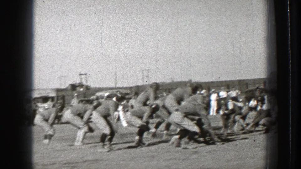 In 1939 Washington footage, helmeted football players line up before the snap while spectators and cars stand beyond the field.