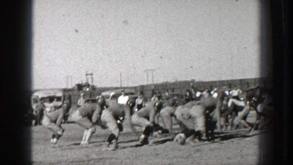 Helmeted players in period uniforms surge from the line on a Washington football field, with spectators and parked cars in the background.
