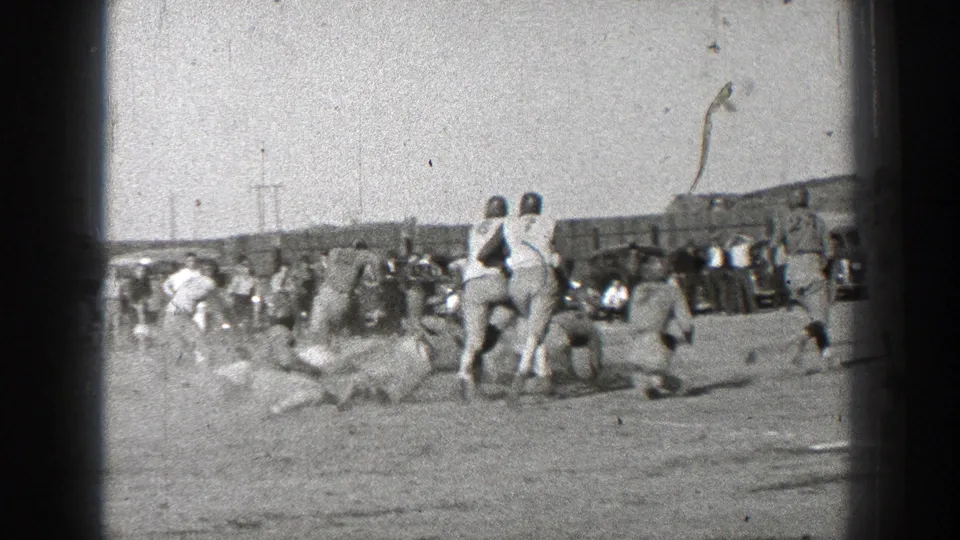 1939 Washington home-movie frame showing helmeted players in motion on a dusty field while spectators watch from the sideline.