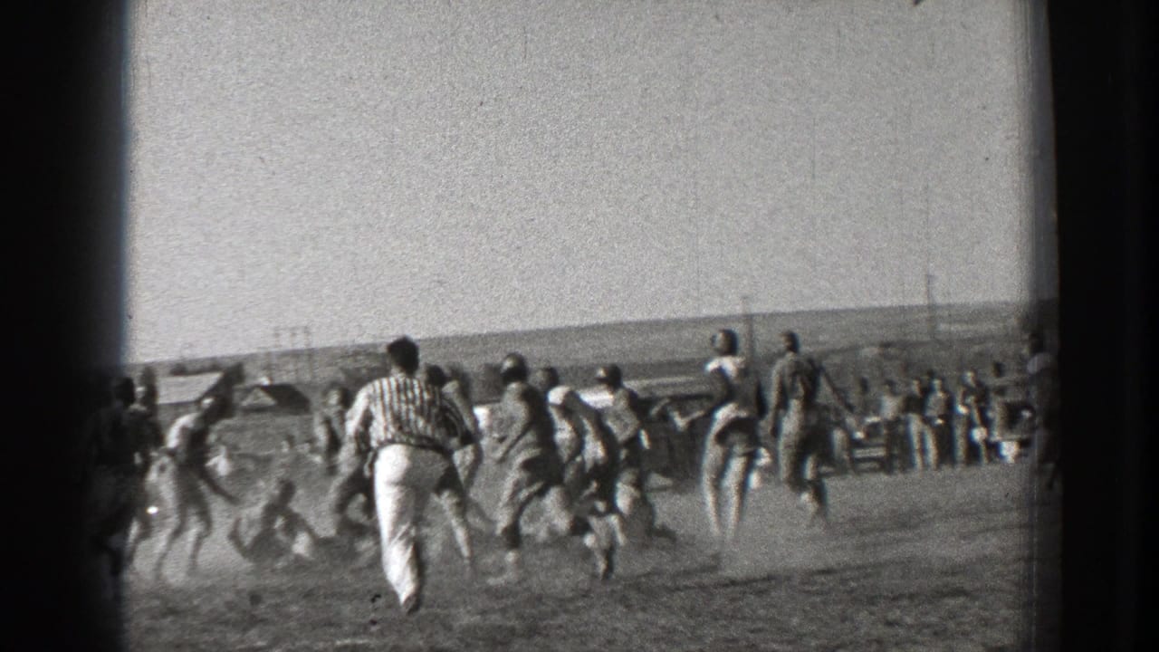 Football players run across a dusty field while spectators watch from the background.