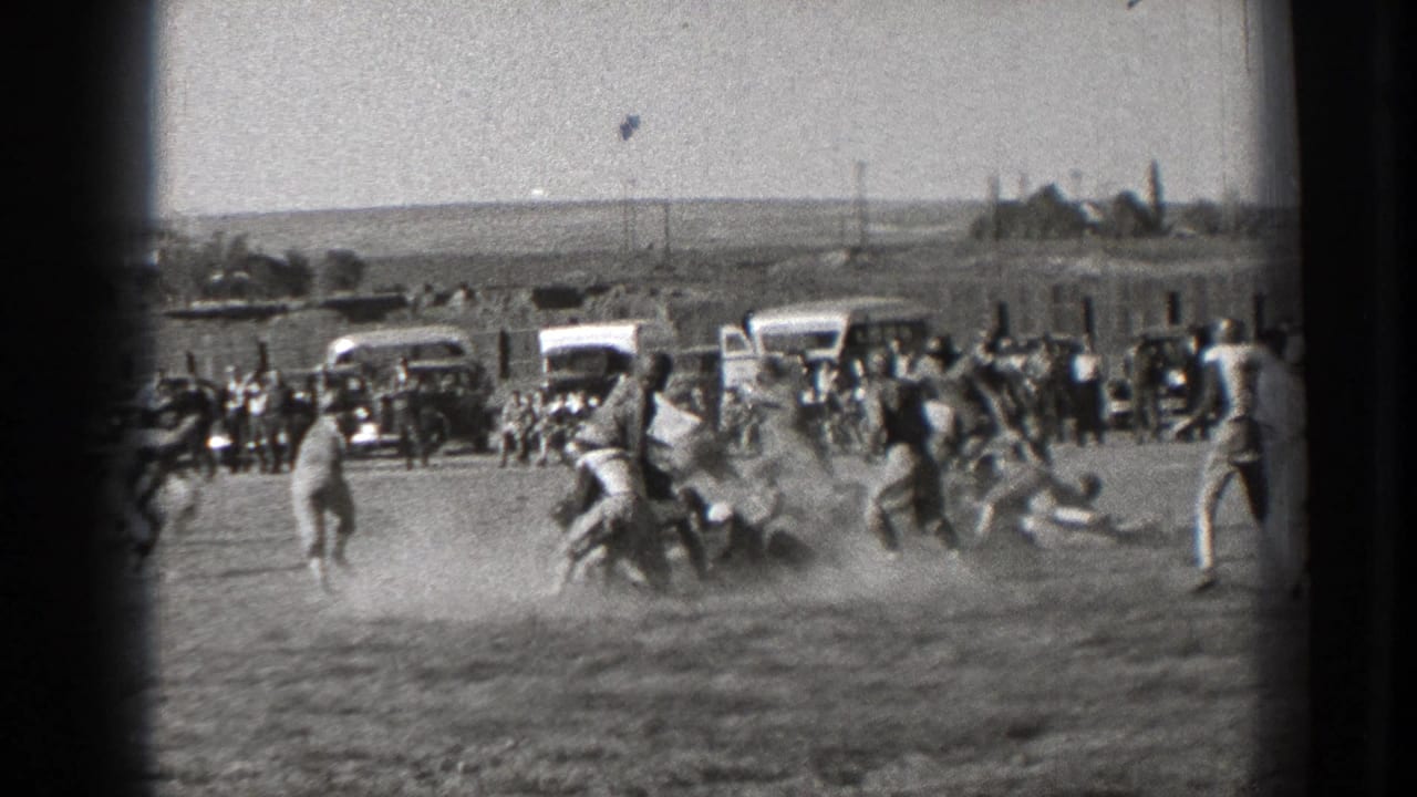 Players collide or tackle on a dusty football field with cars and spectators behind them.