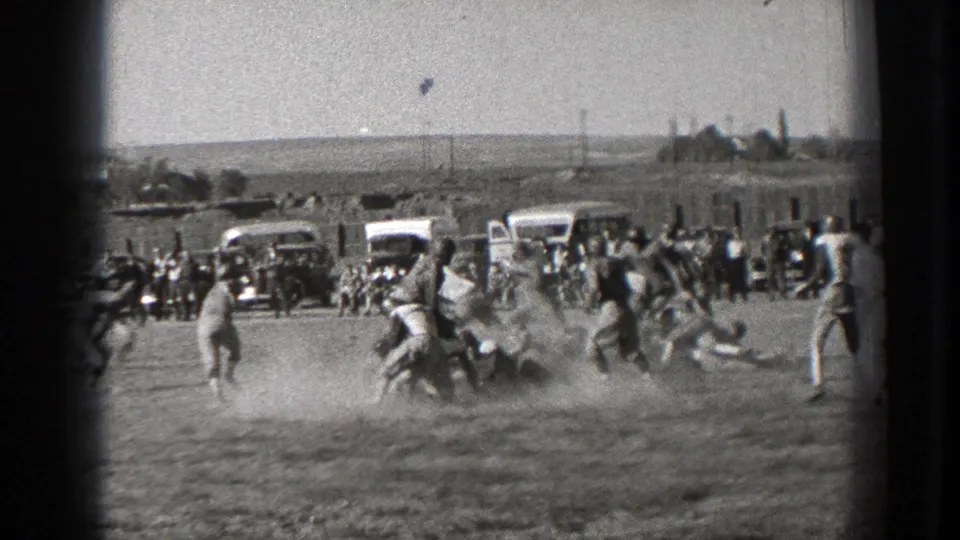 Washington 1939 frame of a football play breaking into a tackle, with dust, parked automobiles, and a crowd along the far sideline.