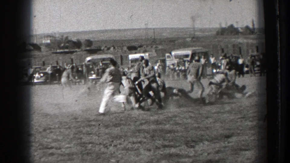 Washington 1939 8mm frame showing players clustered during a football play on a dusty field, with a sideline crowd and older cars behind them.