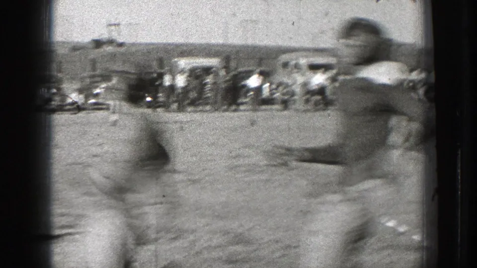 Washington 1939 frame with motion-blurred football players near the camera, a crowd, parked automobiles, and field structures beyond.
