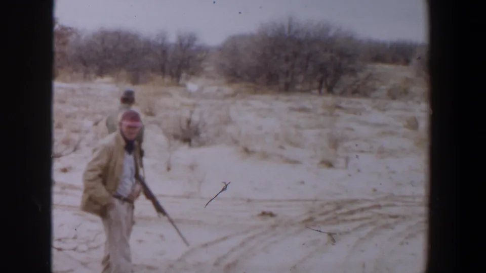 1956 Lubbock, Texas home-movie frame with two adult hunters carrying rifles in a sparse desert-like setting.
