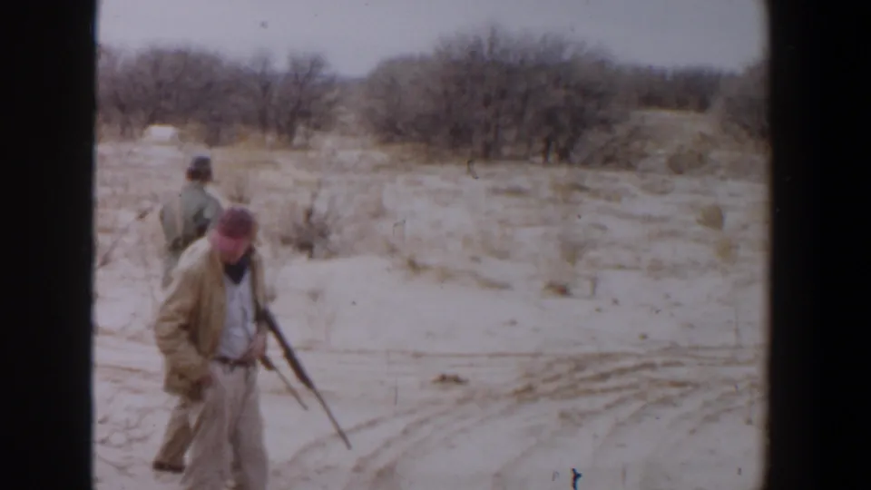 1956 Lubbock, Texas 8mm frame showing hunters with rifles moving through sandy open ground and scrub vegetation.