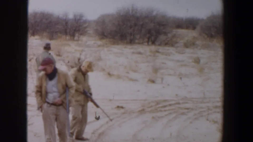 1956 Lubbock, Texas home-movie frame of two men carrying rifles across an arid, brushy landscape.