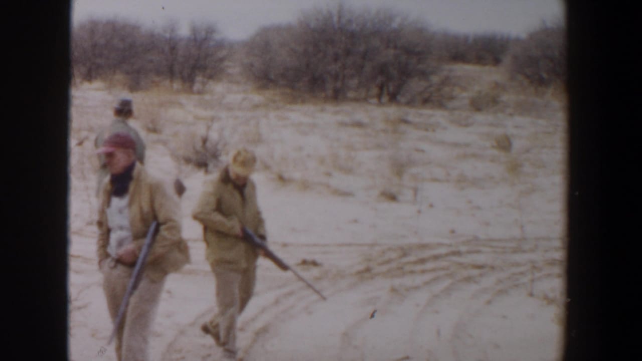 Three men carrying rifles walk across sandy brushland under an overcast sky.