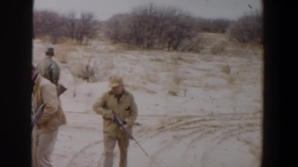 Two adult hunters with rifles pass the camera as another man stands behind them in the Lubbock, Texas brushland scene.