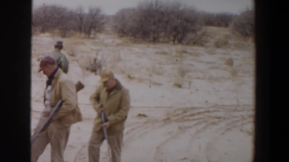 Adult hunters in hats and beige jackets walk close to the camera with rifles in a 1956 Lubbock, Texas home-movie frame.