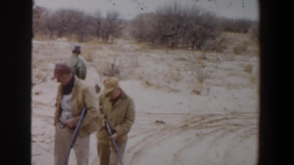 Three adult hunters continue across sandy West Texas terrain with rifles, sparse brush, and leafless trees behind them.